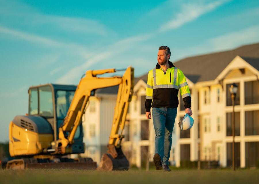 Bearded worker in reflective jacket walks away from heavy equipment at construction site.