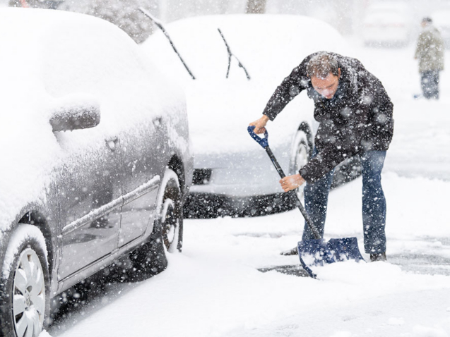 Young man, male in winter coat cleaning, shoveling driveway, street from covered falling, snow in heavy snowing snowstorm with shovel, residential houses, cars parked on road
