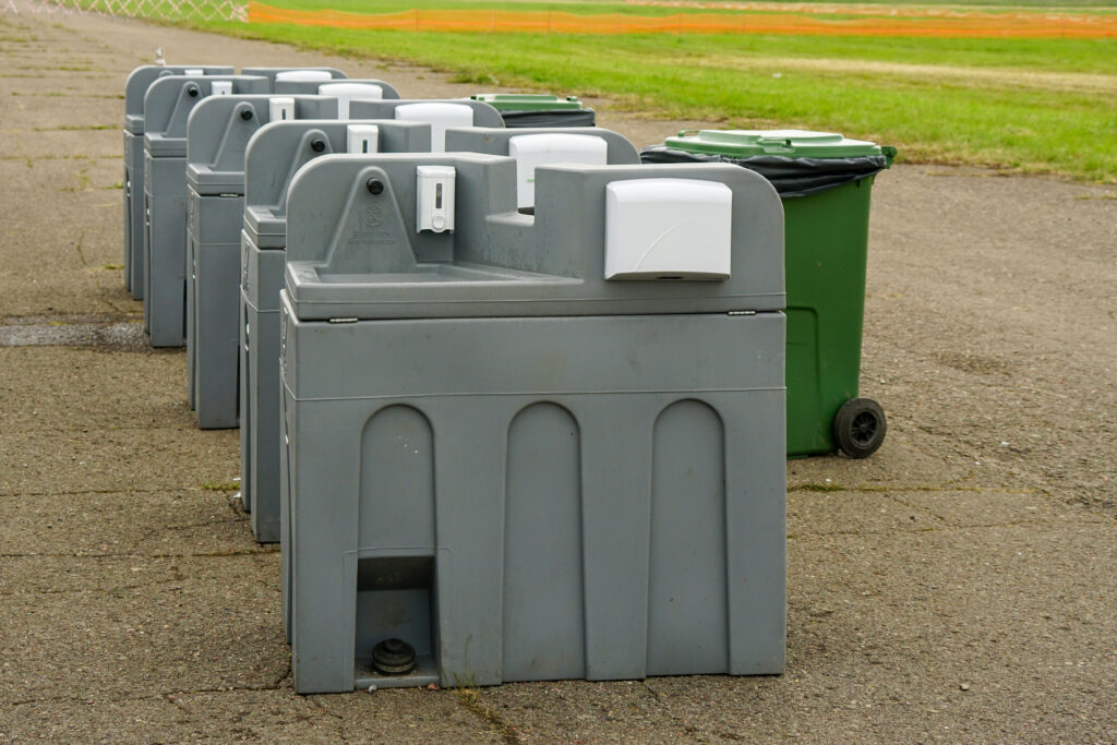 Row of portable outdoor hand wash stations with soap and paper towel dispensers next to a trash bin