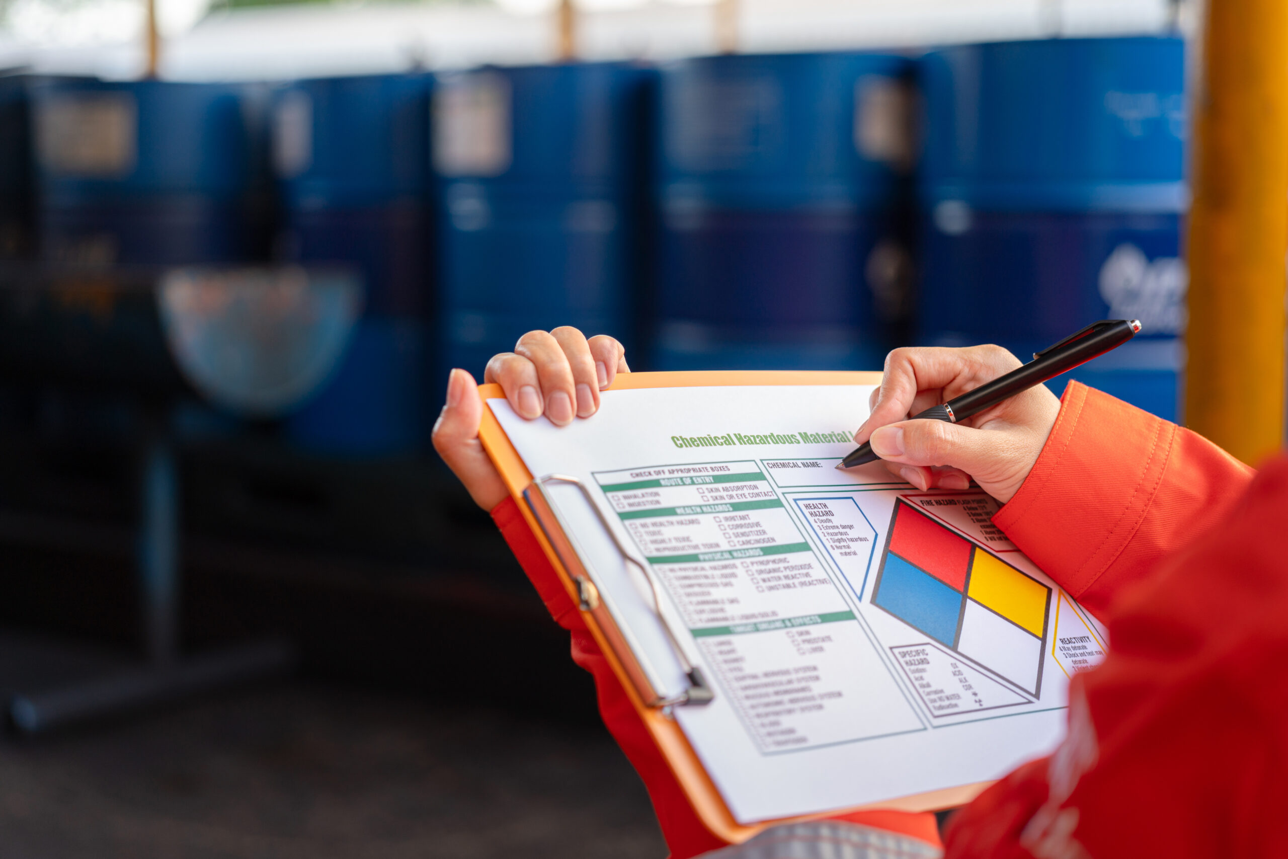 Worker completing chemical hazardous material assessment form on clipboard in front of storage drums