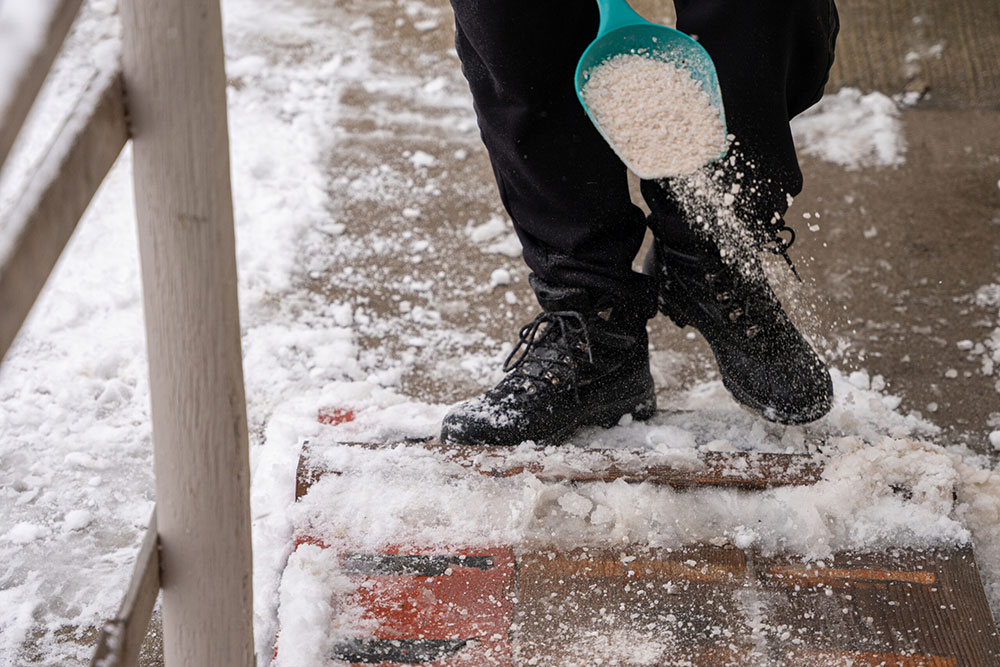 Worker spreading rock salt with scoop to de-ice a snow-covered walkway during winter storm